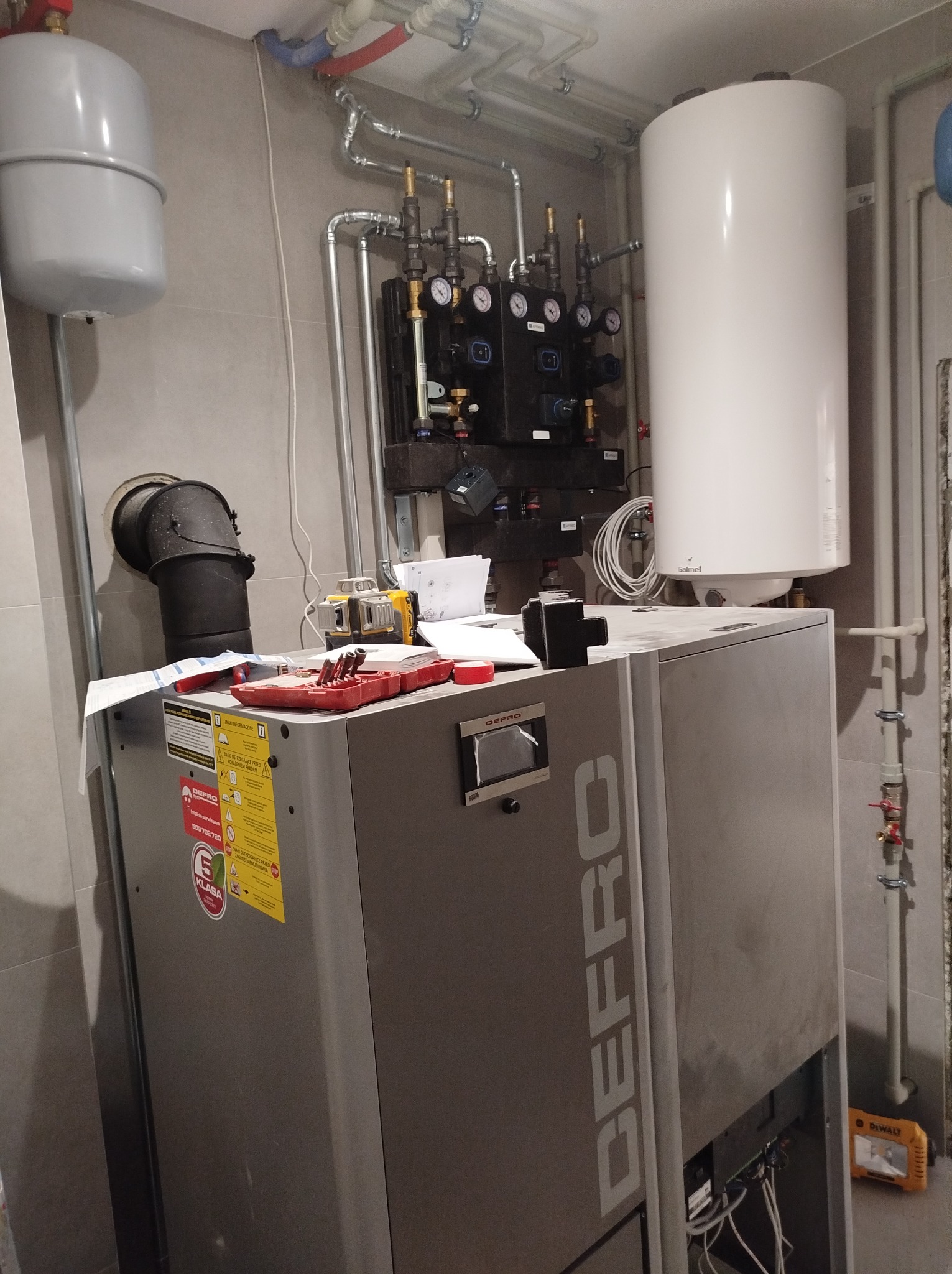 Szary piec DEFRO z podłączoną instalacją grzewczą i zasobnikiem ciepłej wody, narzędzia na piecu, widoczne rury instalacji CO i CWU.