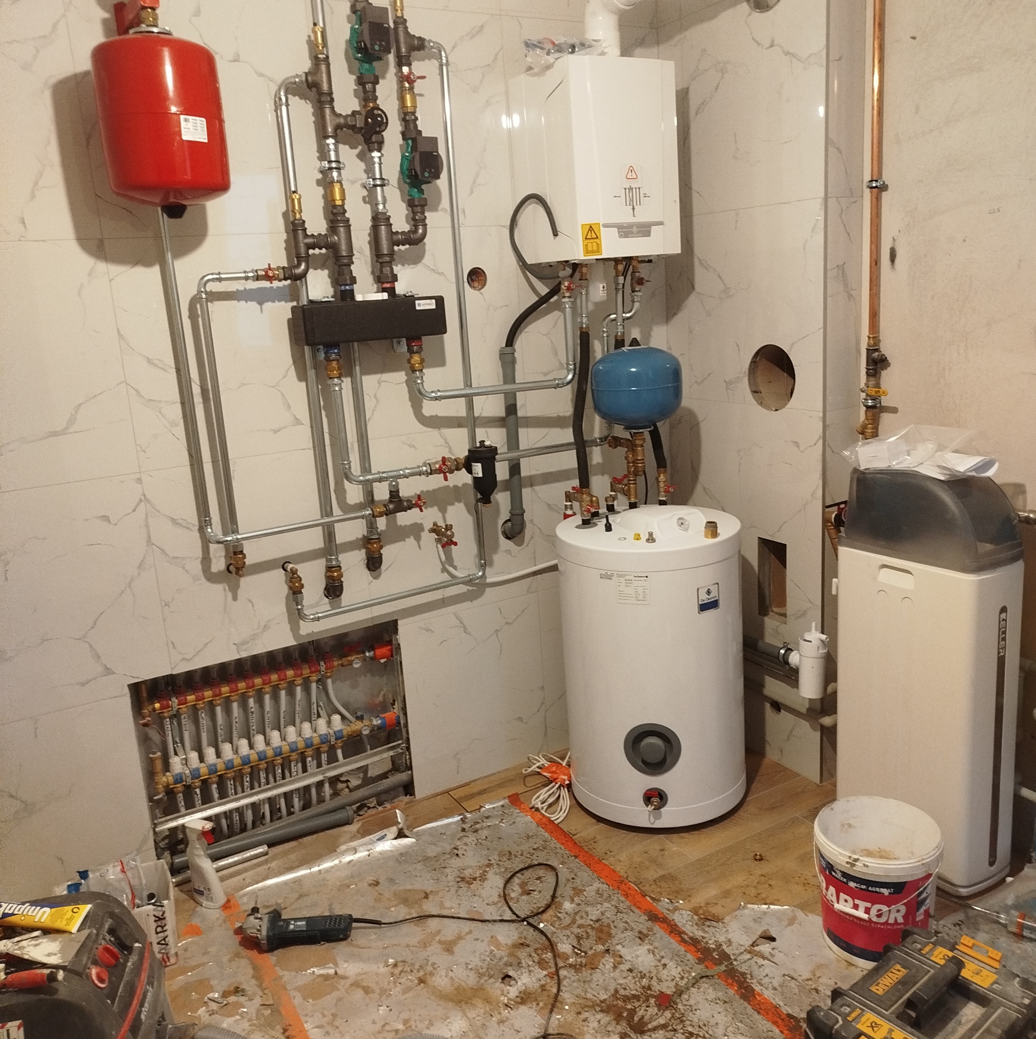 Wnętrze kotłowni w trakcie montażu: widoczny rozdzielacz ogrzewania podłogowego, zasobnik ciepłej wody z niebieskim naczyniem przeponowym, piec gazowy, zmiękczacz wody Keller, oraz narzędzia...