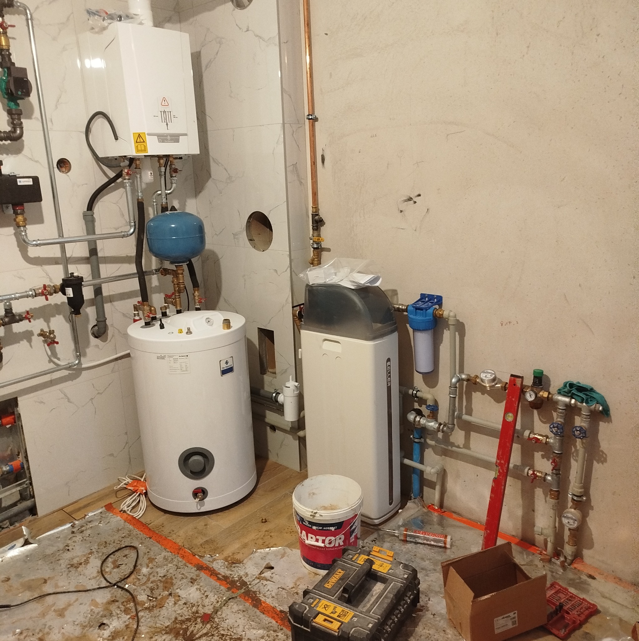 Wnętrze w trakcie instalacji: bojler, piec gazowy, zmiękczacz wody oraz rury miedziane i plastikowe, widoczne narzędzia i materiały budowlane na podłodze.