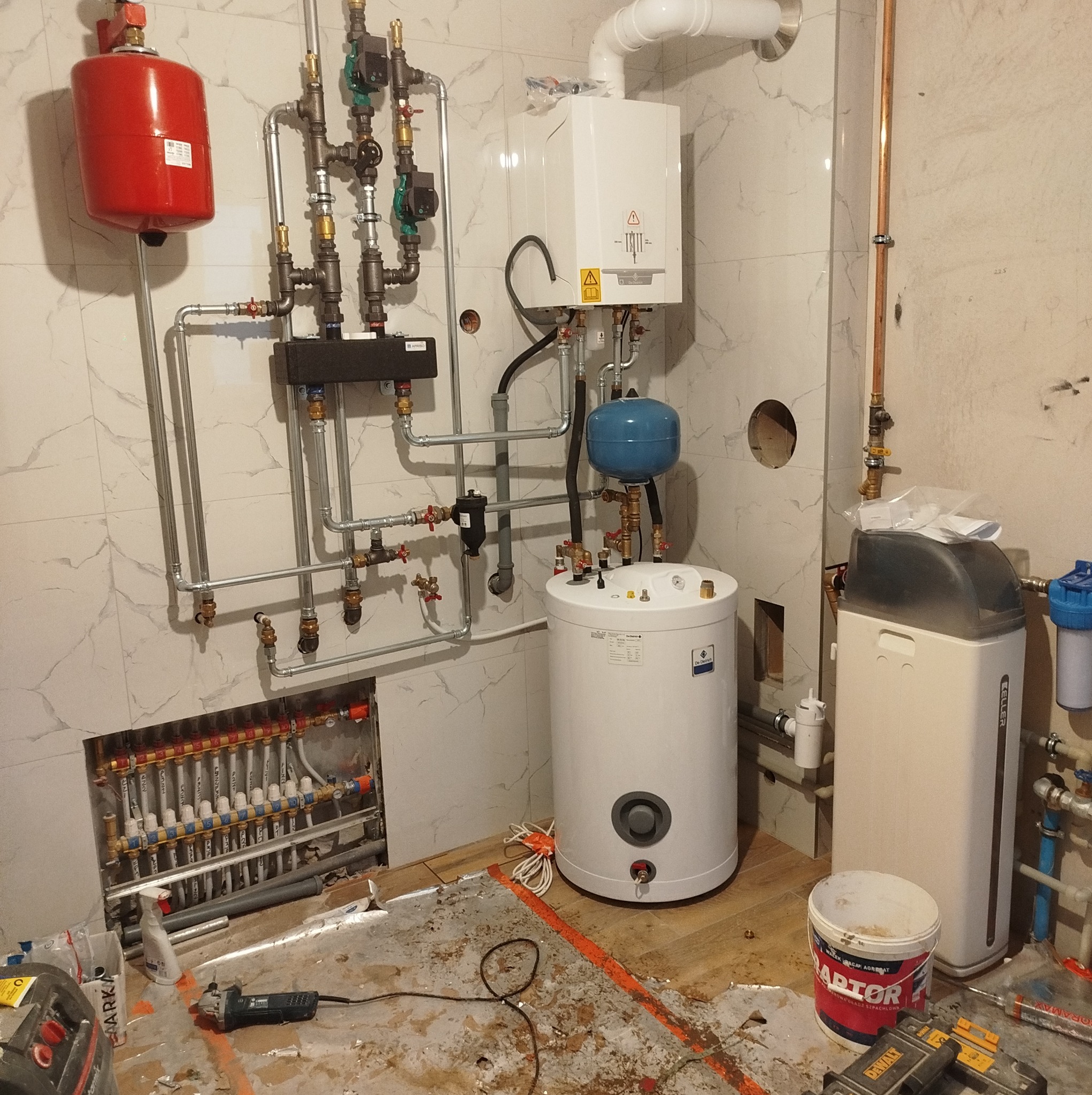 Wnętrze łazienki w trakcie prac instalacyjnych, widoczny rozdzielacz ogrzewania podłogowego, piec gazowy, zasobnik ciepłej wody, zmiękczacz wody, liczne rury i zawory, oraz narzędzia i bałagan...