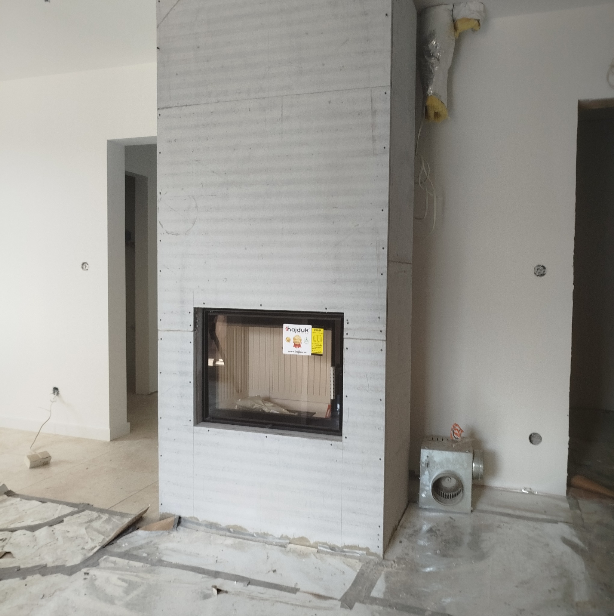 Wnętrze w trakcie prac wykończeniowych z widocznym obudowanym kominkiem z wkładem, wentylatorem i zabezpieczoną podłogą.
