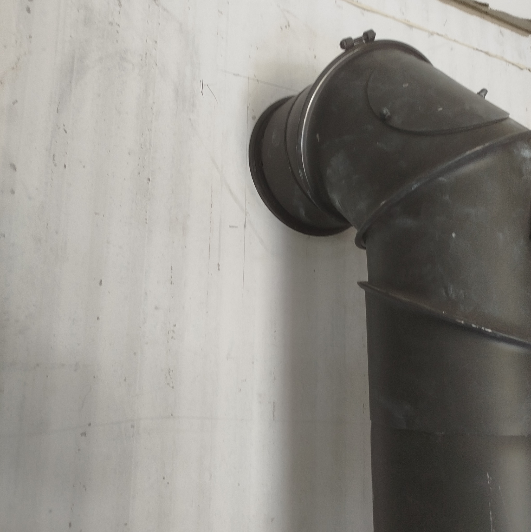 Czarny, metalowy przewód kominowy zamontowany do szarej, surowej ściany w Mońkach, widoczne ślady po ołówku i niedoskonałości tynku.