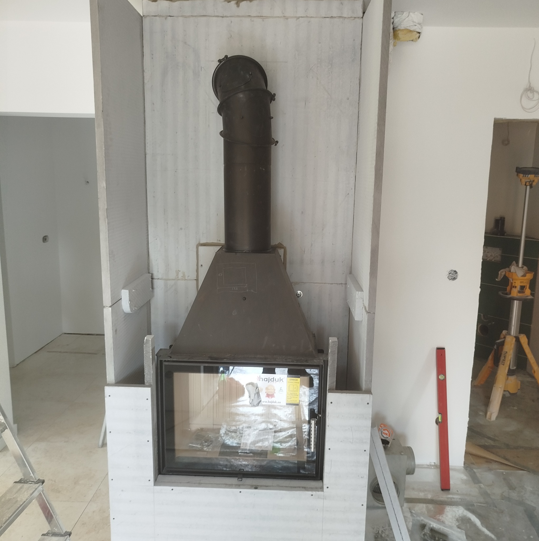 Wnętrze w trakcie wykańczania z zamontowanym wkładem kominkowym Hajduk, obudowanym płytami gipsowymi, widoczny przewód dymowy i poziomica oparta o ścianę.