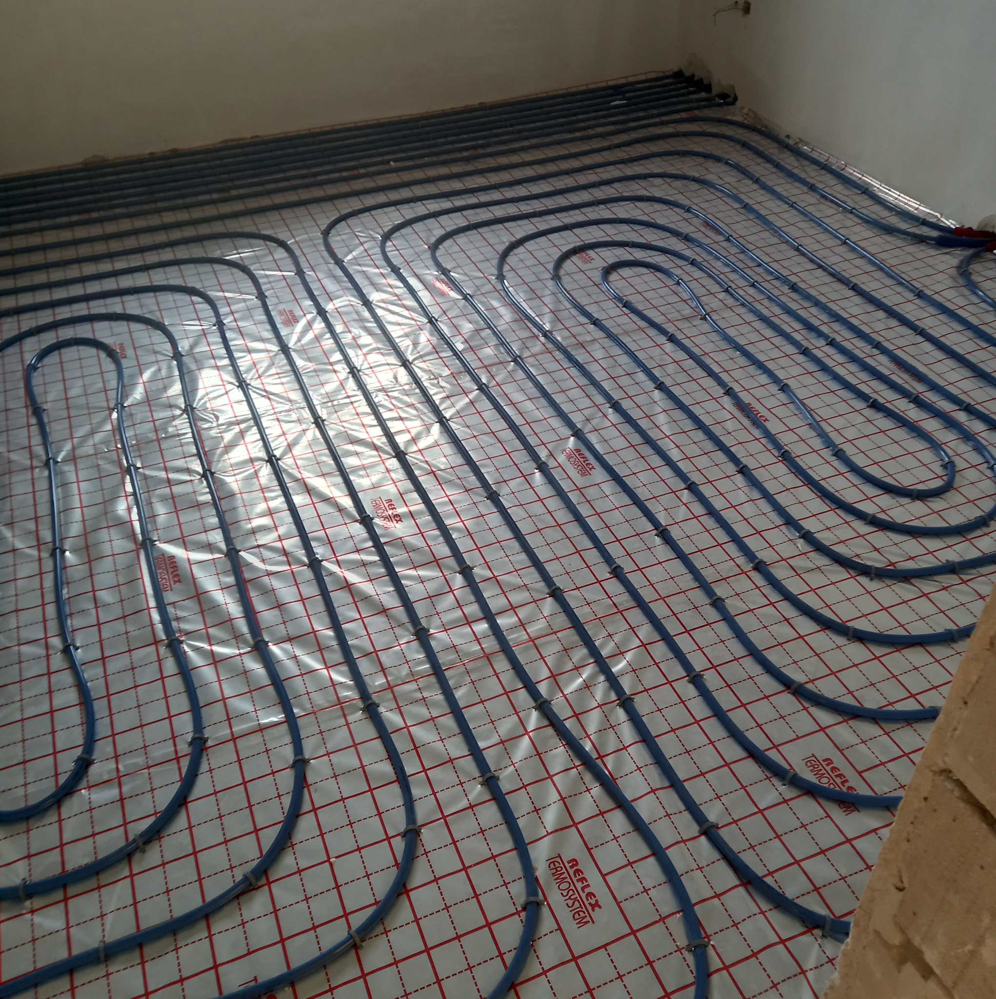 Niebieskie rury ogrzewania podłogowego ułożone na folii z czerwoną siatką, widok z góry, fragment ściany z prawej strony.