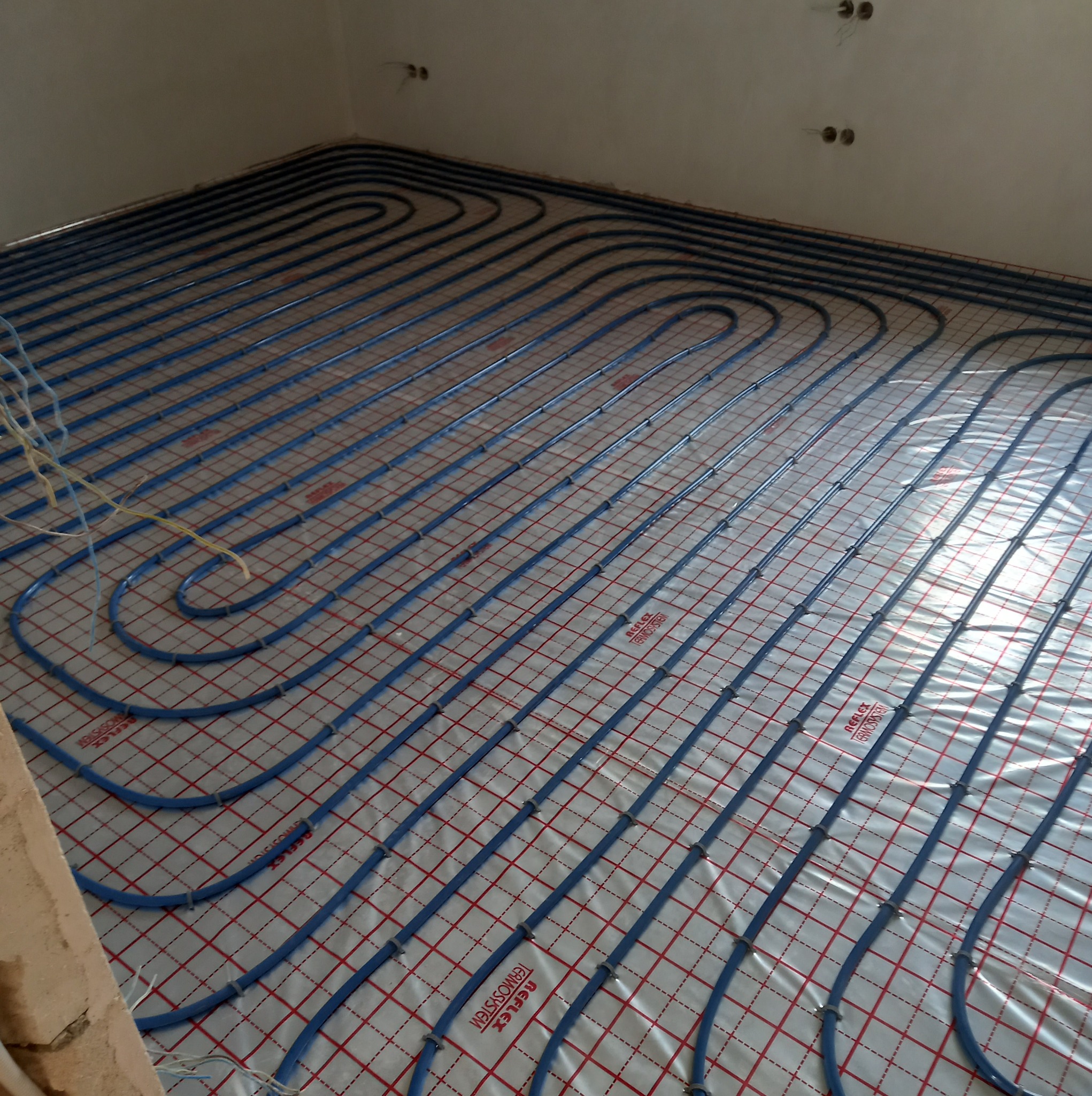 Niebieskie rury systemu ogrzewania podłogowego ułożone na folii z czerwoną siatką w surowym wnętrzu, widoczne kable elektryczne i niedokończona ściana.