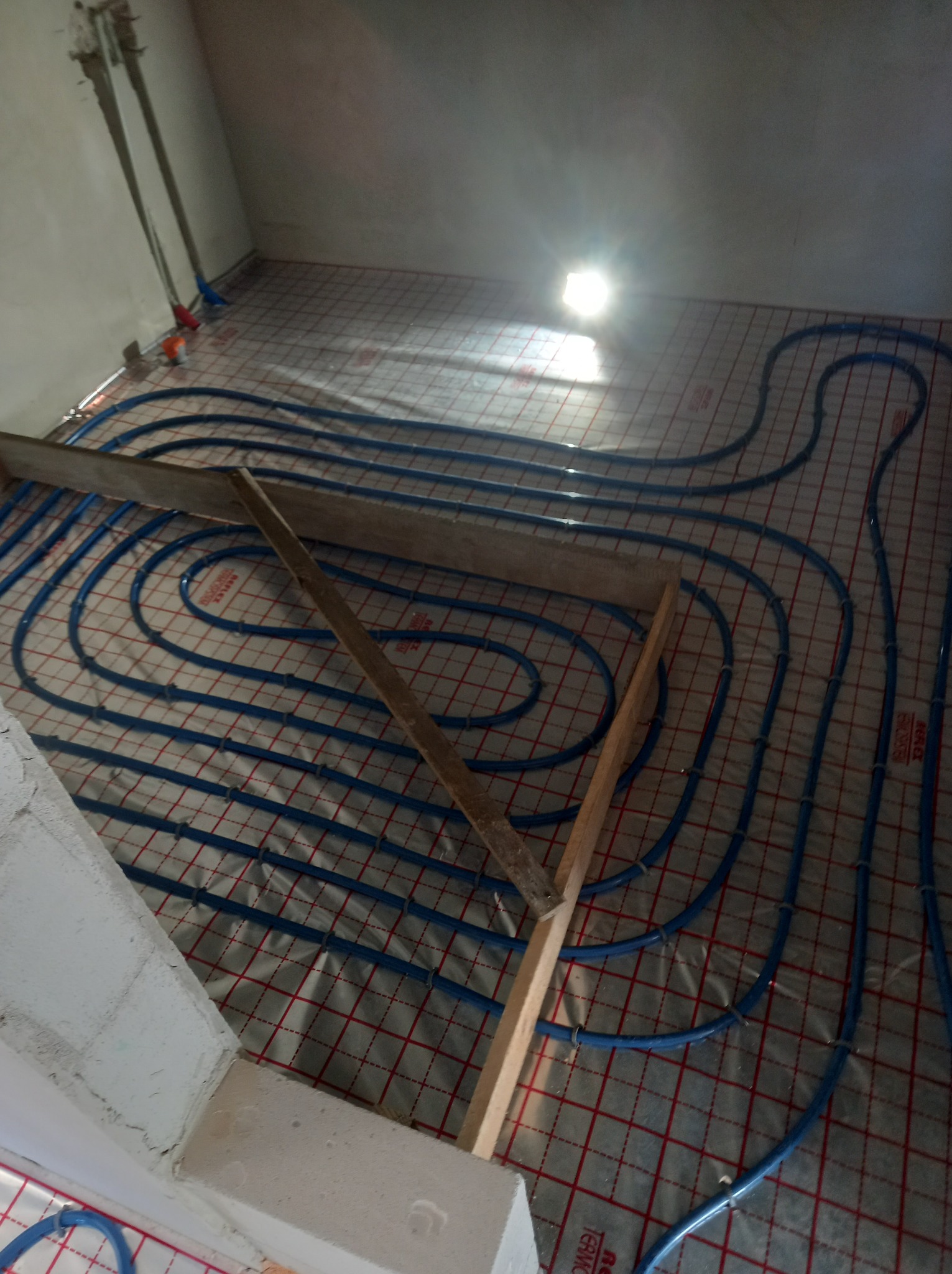 Niedokończona instalacja ogrzewania podłogowego z niebieskimi rurami na folii z czerwoną siatką, tymczasowo zabezpieczona drewnianymi elementami, widok z góry w surowym wnętrzu.