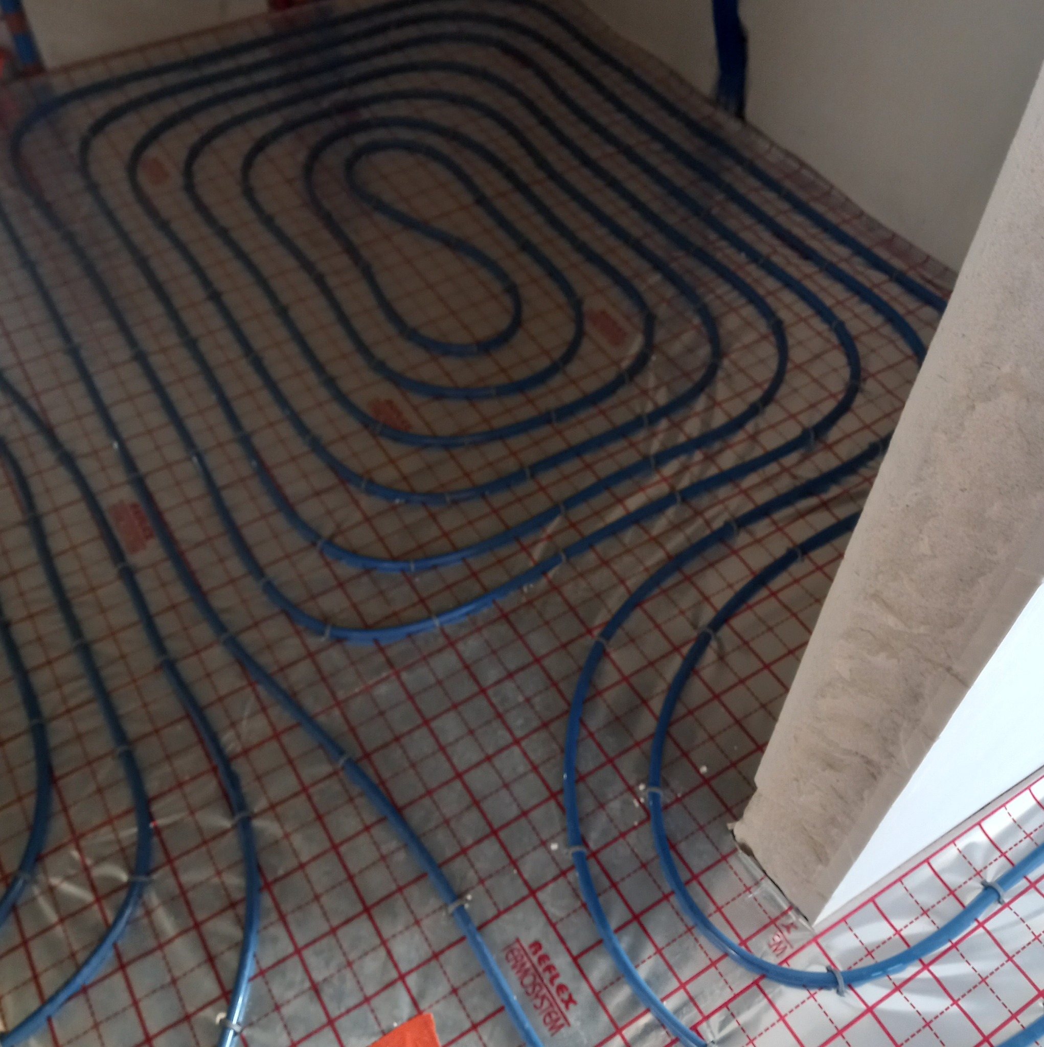 Niebieskie rurki systemu ogrzewania podłogowego ułożone na folii refleksyjnej z czerwoną siatką, widoczne mocowania rur do podłoża i fragment surowego narożnika ściany.