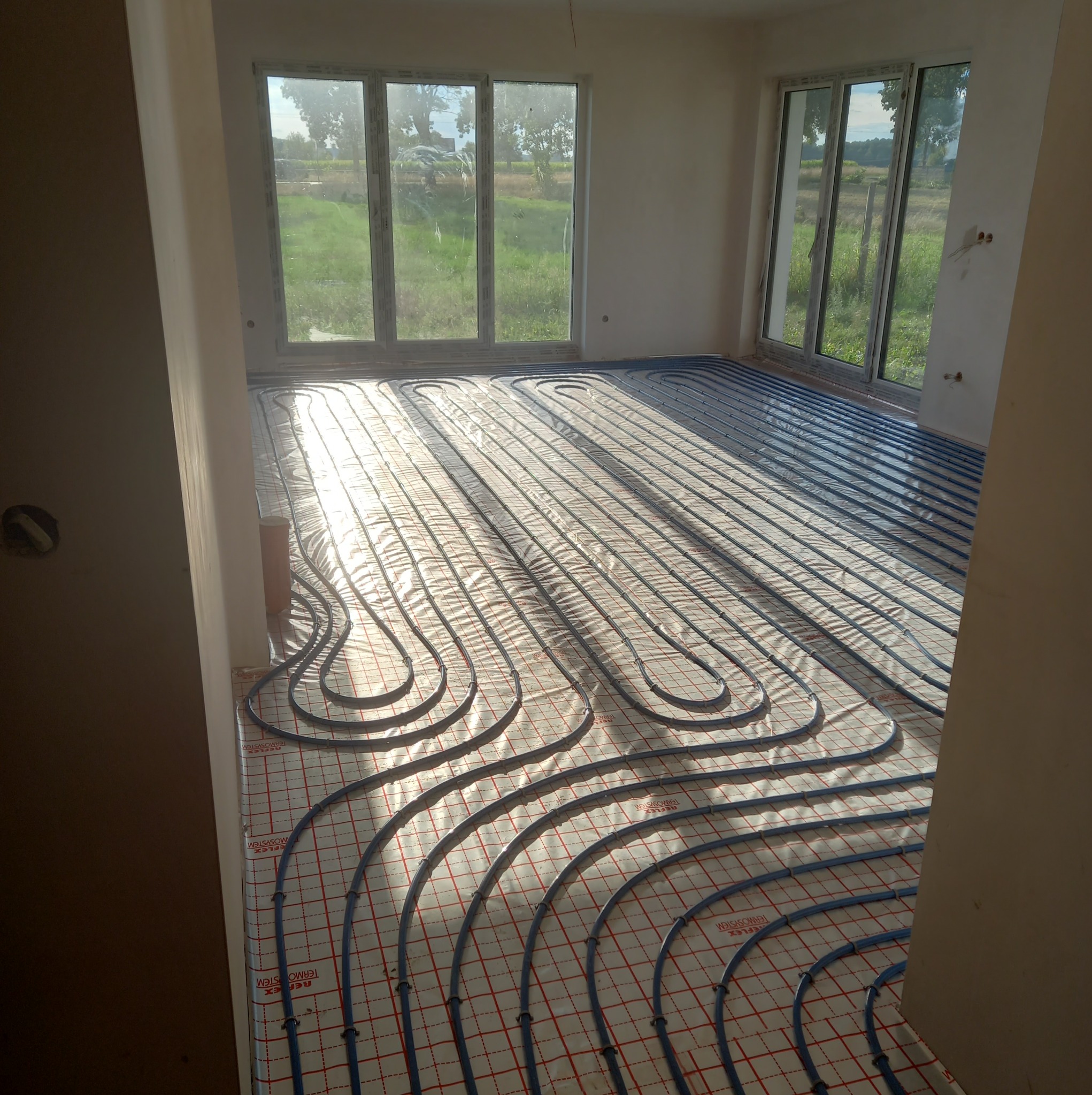 Niebieskie rury instalacji grzewczej podłogowej ułożone na macie izolacyjnej z siatką w nowym budynku, widok zza futryny drzwi, w tle okno z widokiem na zieleń.