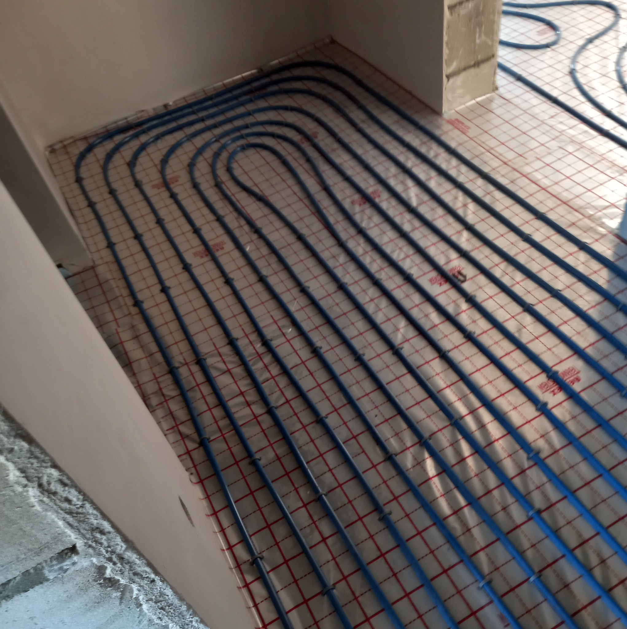 Błękitne rury systemu ogrzewania podłogowego ułożone w pętle na folii z czerwoną kratką w surowym wnętrzu, widok z góry pod kątem.