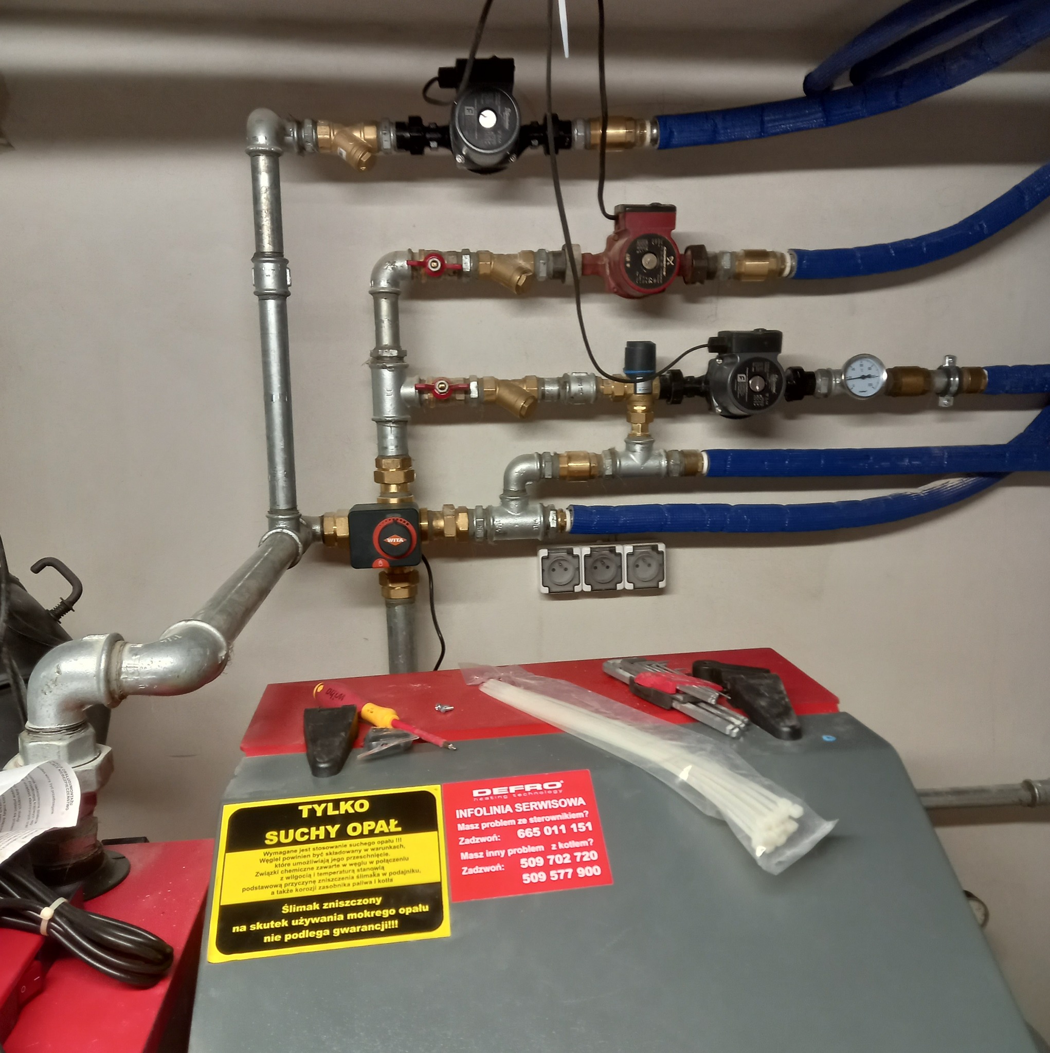 Instalacja grzewcza z rurami, pompami obiegowymi i zaworami, widoczne narzędzia i etykieta serwisowa Defro na kotle.