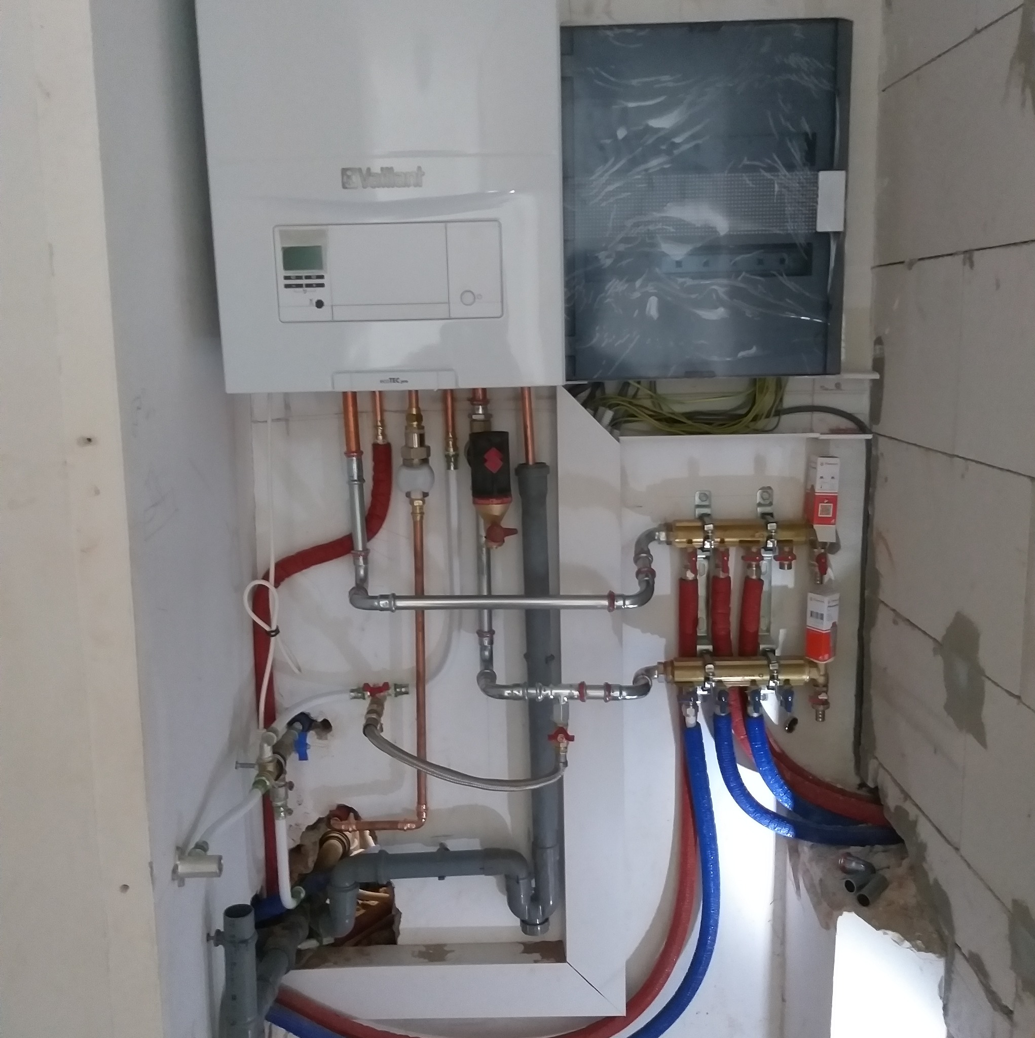 Nowoczesny piec gazowy Vaillant z podłączeniem do instalacji centralnego ogrzewania i rozdzielaczem ogrzewania podłogowego, widoczne rury miedziane i PEX w kolorach czerwonym i niebieskim, ściany...