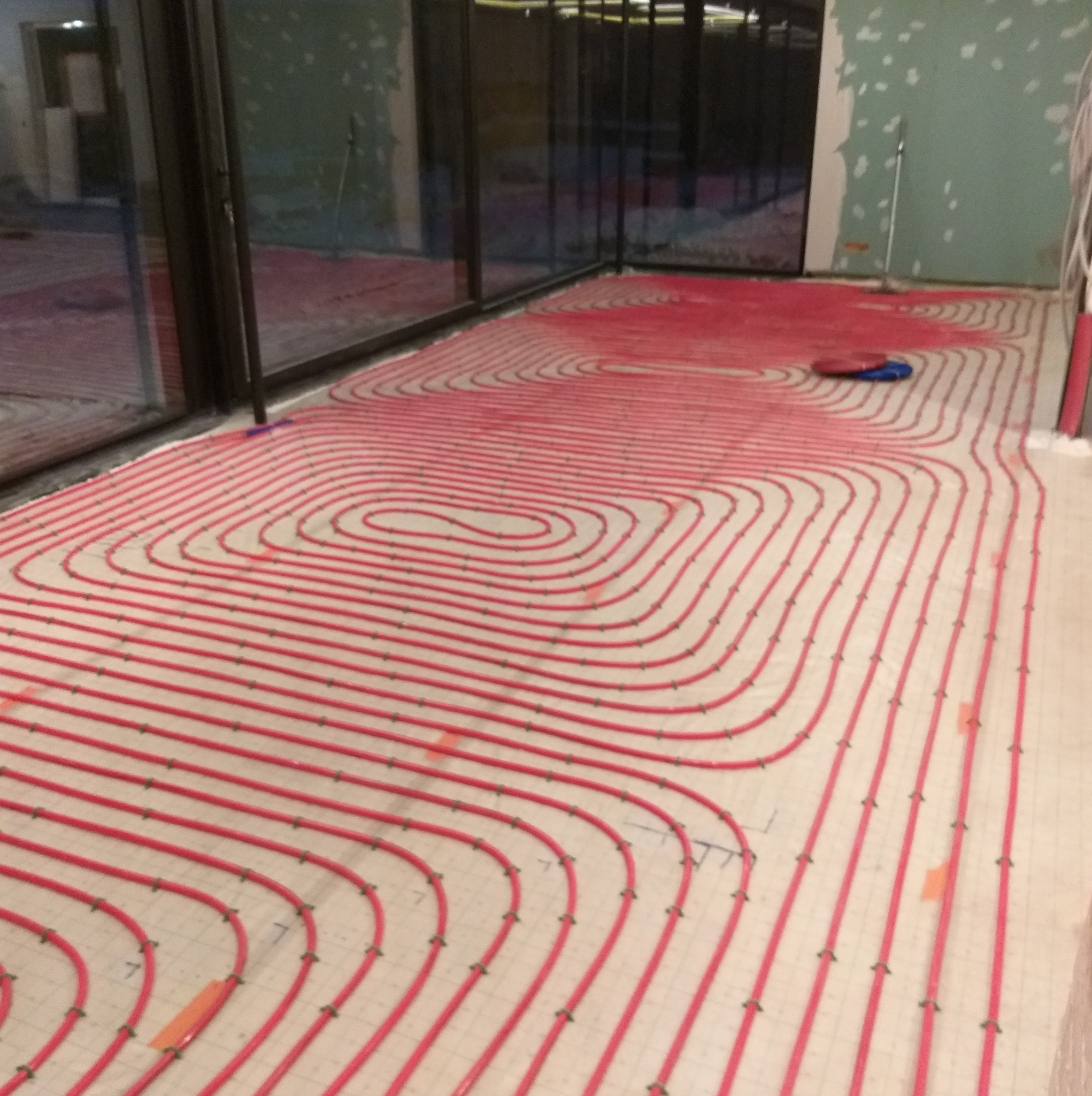 Czerwone rurki ogrzewania podłogowego ułożone spiralnie na białej macie, widoczne mocowania i fragmenty taśmy, surowy stan wykończenia pomieszczenia z dużym oknem.