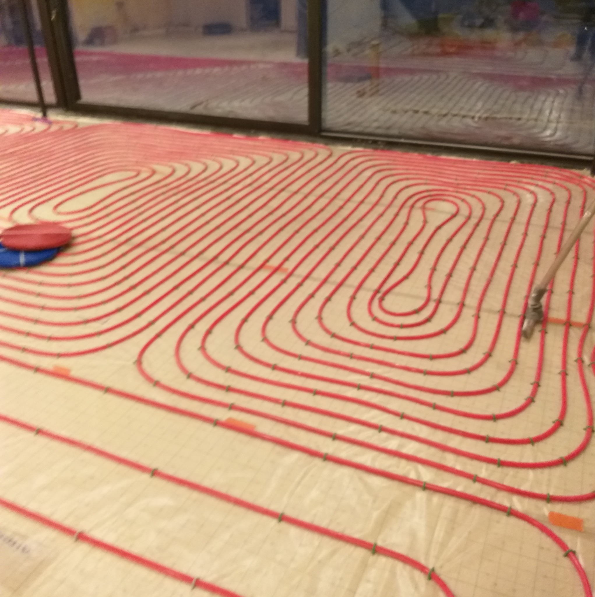 Czerwone rury układane w pętle na podłożu z siatką, tworzące system ogrzewania podłogowego, widoczne zielone spinki mocujące, w tle przesuwne okno.