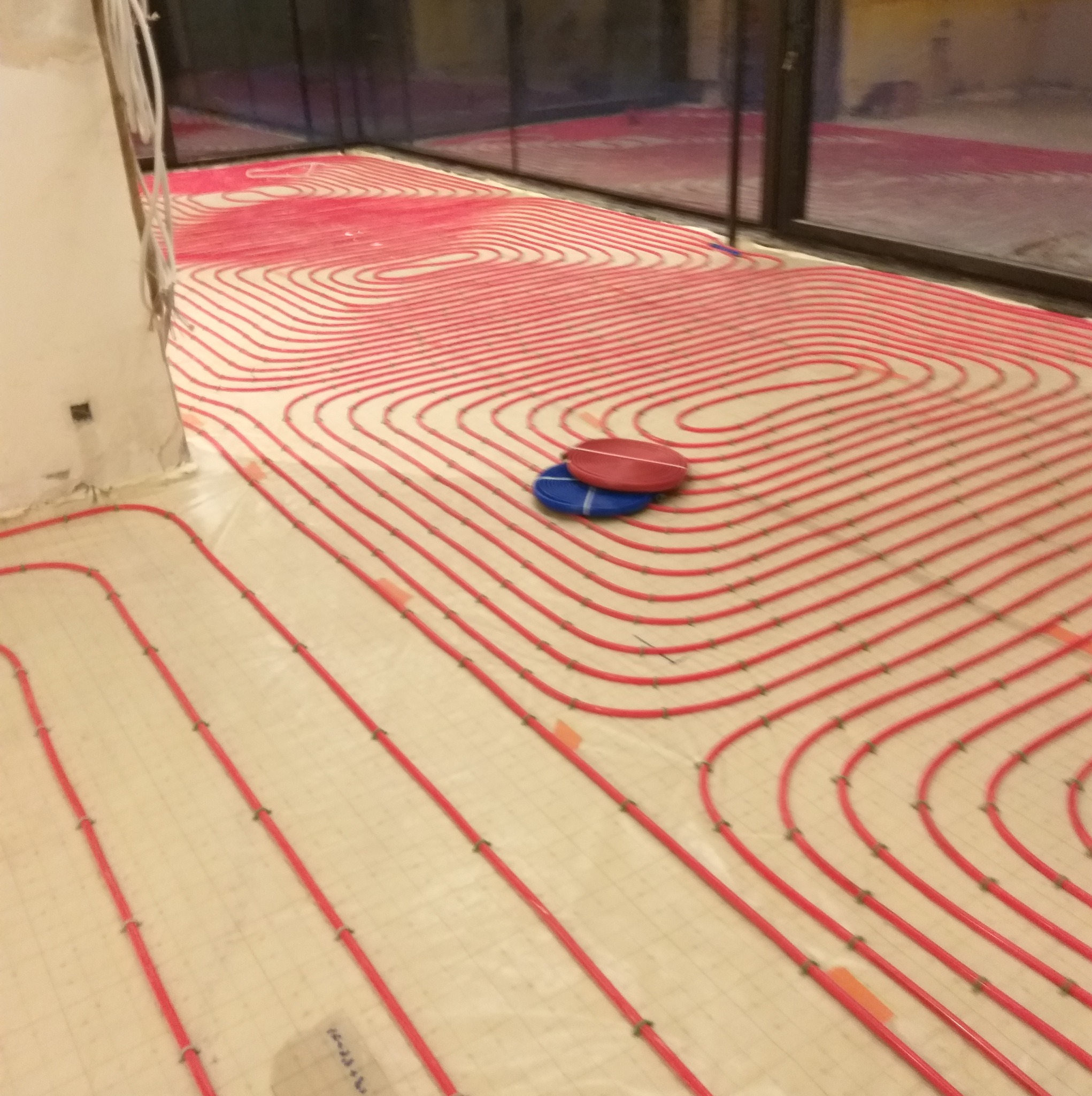 Czerwone rurki systemu grzewczego podłogowego ułożone spiralnie na białej macie izolacyjnej, widoczne łączniki i dwa krążki taśmy: czerwony i niebieski.