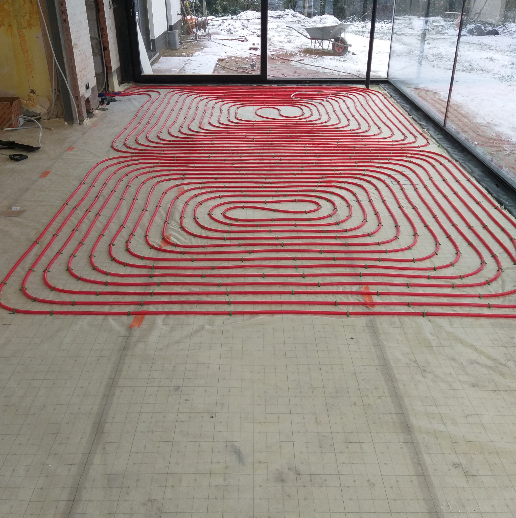 Czerwone rurki systemu rozprowadzenia ciepła, przymocowane zielonymi uchwytami do folii z nadrukowaną siatką, w surowym wnętrzu z widokiem na ośnieżony ogród.