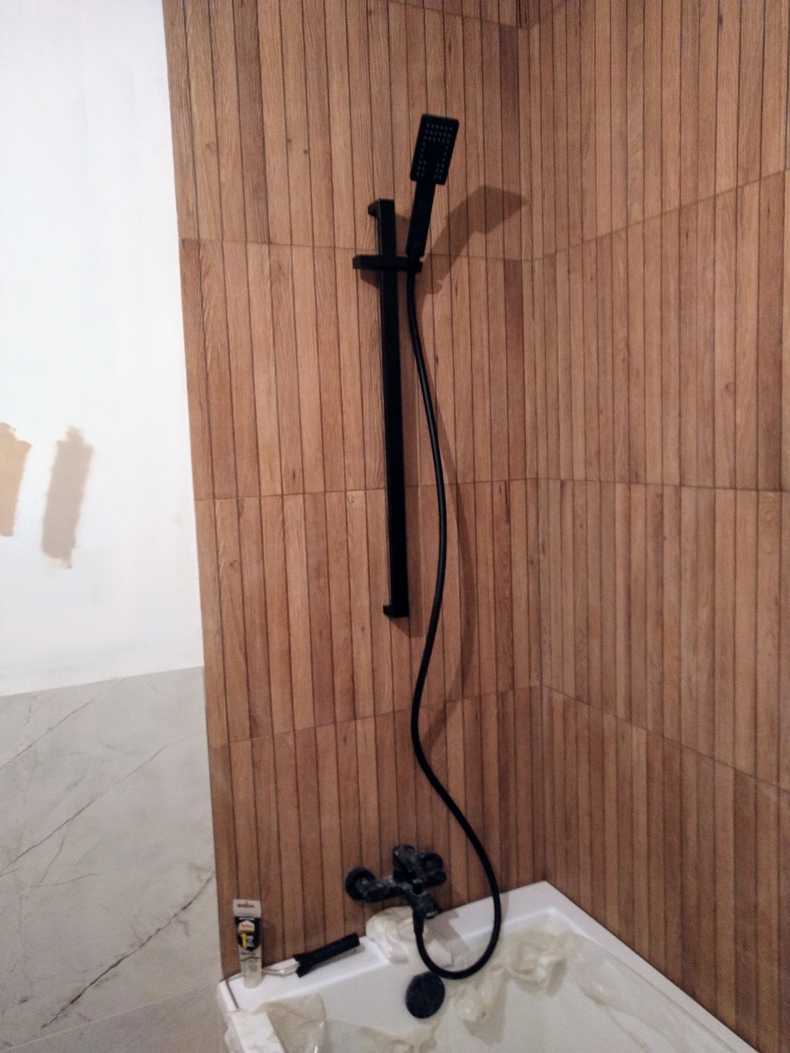 Czarna armatura prysznicowa zamontowana na ścianie wyłożonej płytkami imitującymi drewno w trakcie prac wykończeniowych łazienki, widoczna folia zabezpieczająca wannę i narzędzia.