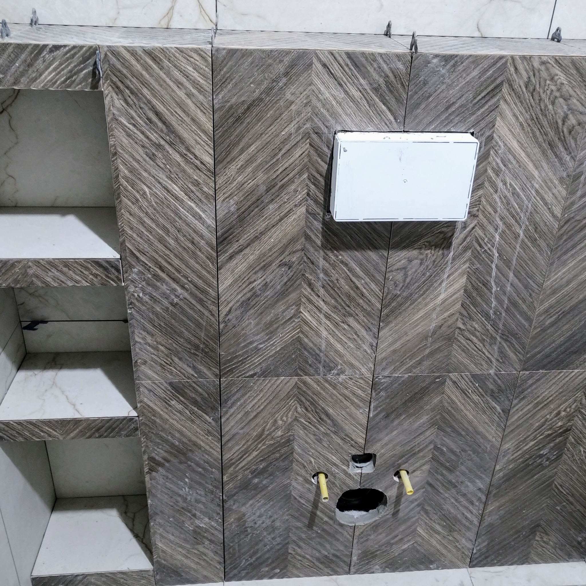 Ściana łazienki w trakcie wykańczania, z ułożonymi płytkami w jodełkę o szarym odcieniu imitującym drewno, widoczna biała obudowa stelaża podtynkowego i przygotowane otwory na instalację sanitarną...