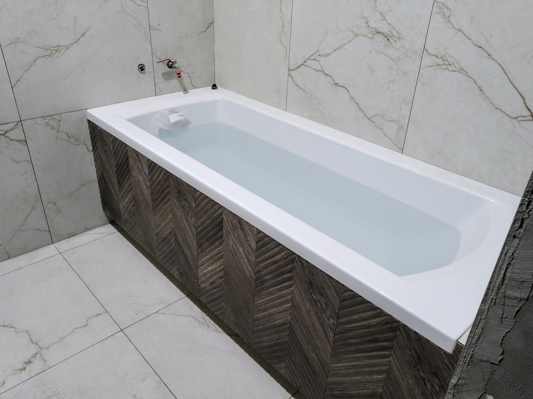 Wanna wypełniona wodą, obudowana panelami w jodełkę, na tle marmurowych płytek ściennych i podłogowych, z widoczną instalacją wodną.
