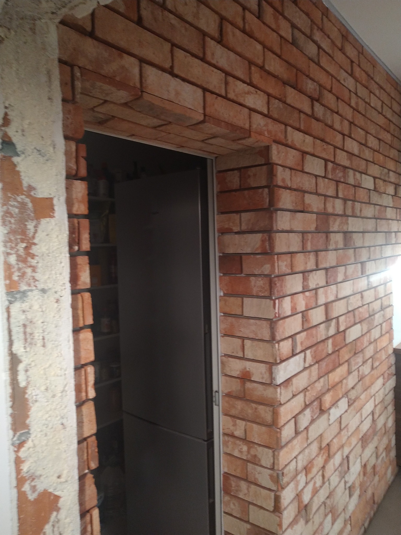 Wykończona ściana z cegły klinkierowej wewnątrz pomieszczenia, widoczne ościeże drzwi w trakcie prac wykończeniowych, z resztkami zaprawy