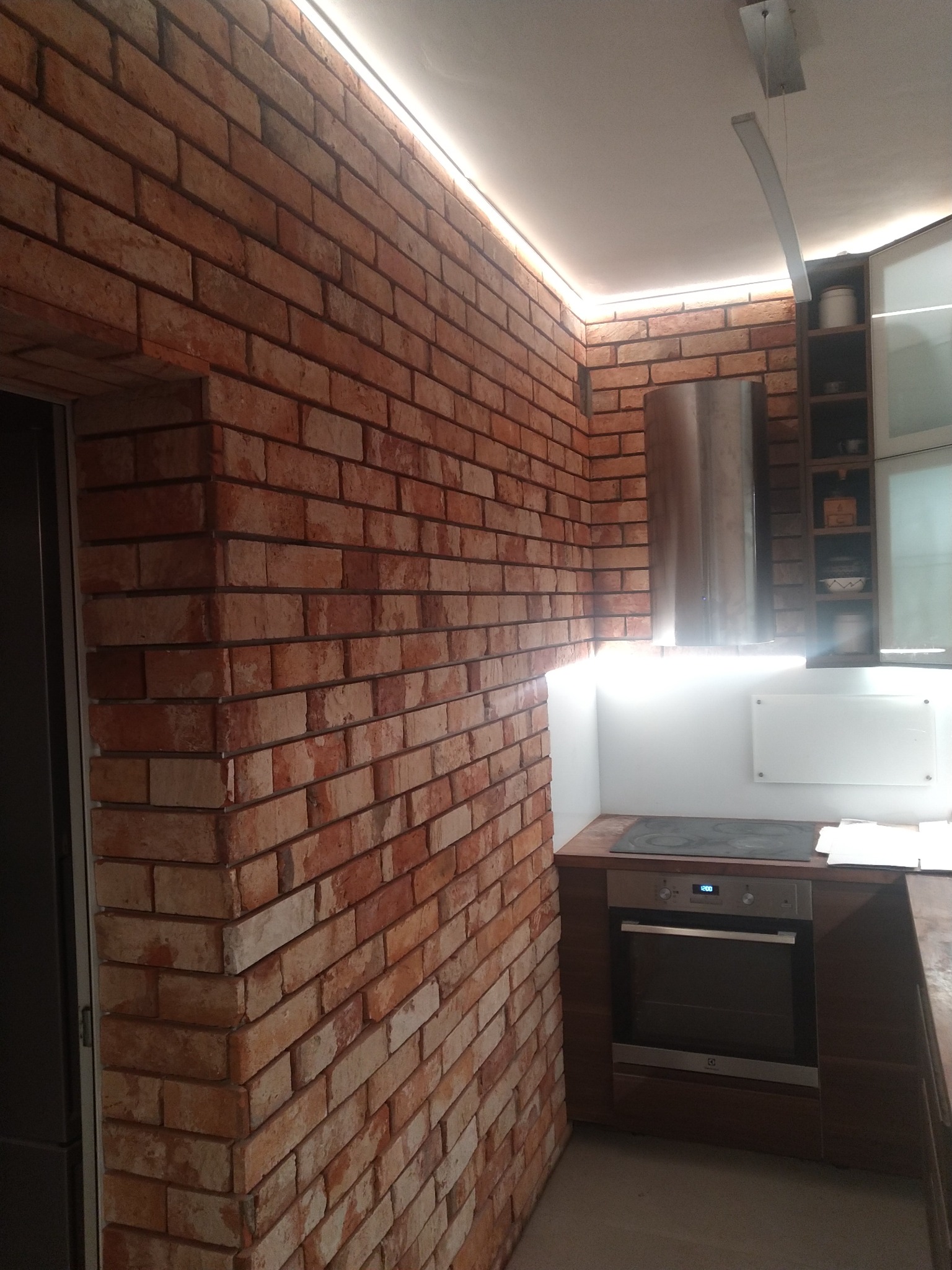 Kuchnia z ceglaną ścianą, oświetleniem LED podsufitowym i nowoczesnym sprzętem AGD, w tym piekarnikiem marki Electrolux.
