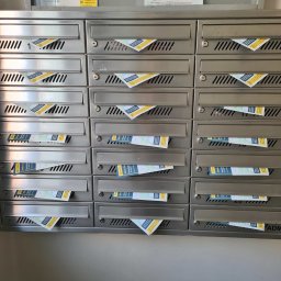 ADRIANET - Rząd metalowych skrzynek pocztowych wypełnionych ulotkami informującymi o konsultacjach dotyczących stref czystego transportu w Krakowie.