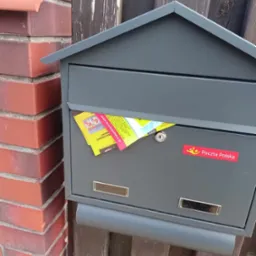Szara skrzynka pocztowa z włożoną ulotką reklamową firmy z Koszalina, naklejka Poczty Polskiej na tle ceglanego muru i drewnianego płotu.