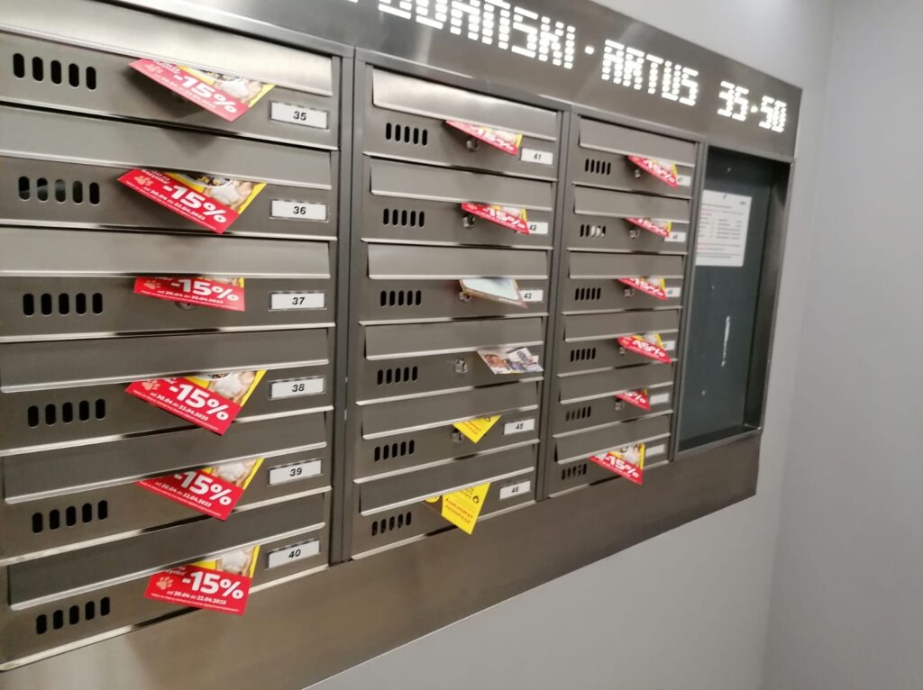 Rząd metalowych skrzynek pocztowych z numerami od 35 do 50, w niektórych skrzynkach włożone ulotki, a na niektórych naklejone czerwone ulotki z informacją o 15% zniżce.