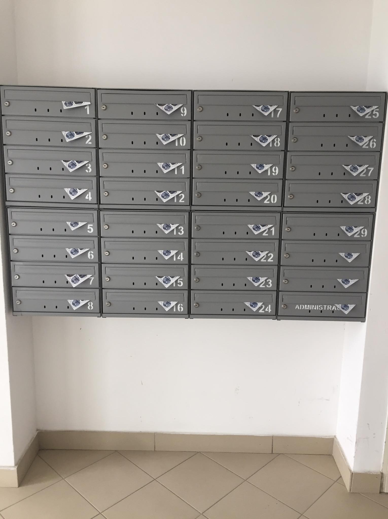 Szare skrzynki pocztowe w bloku mieszkalnym z numerami od 1 do 29 oraz napisem ADMINISTRACJA, niektóre z naklejkami.