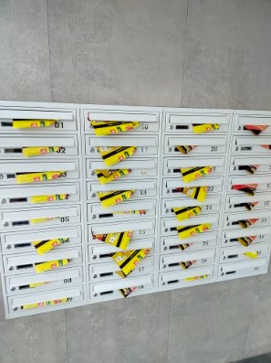 Białe skrzynki pocztowe w bloku, wypełnione żółtymi ulotkami reklamowymi, widoczne numery skrzynek.