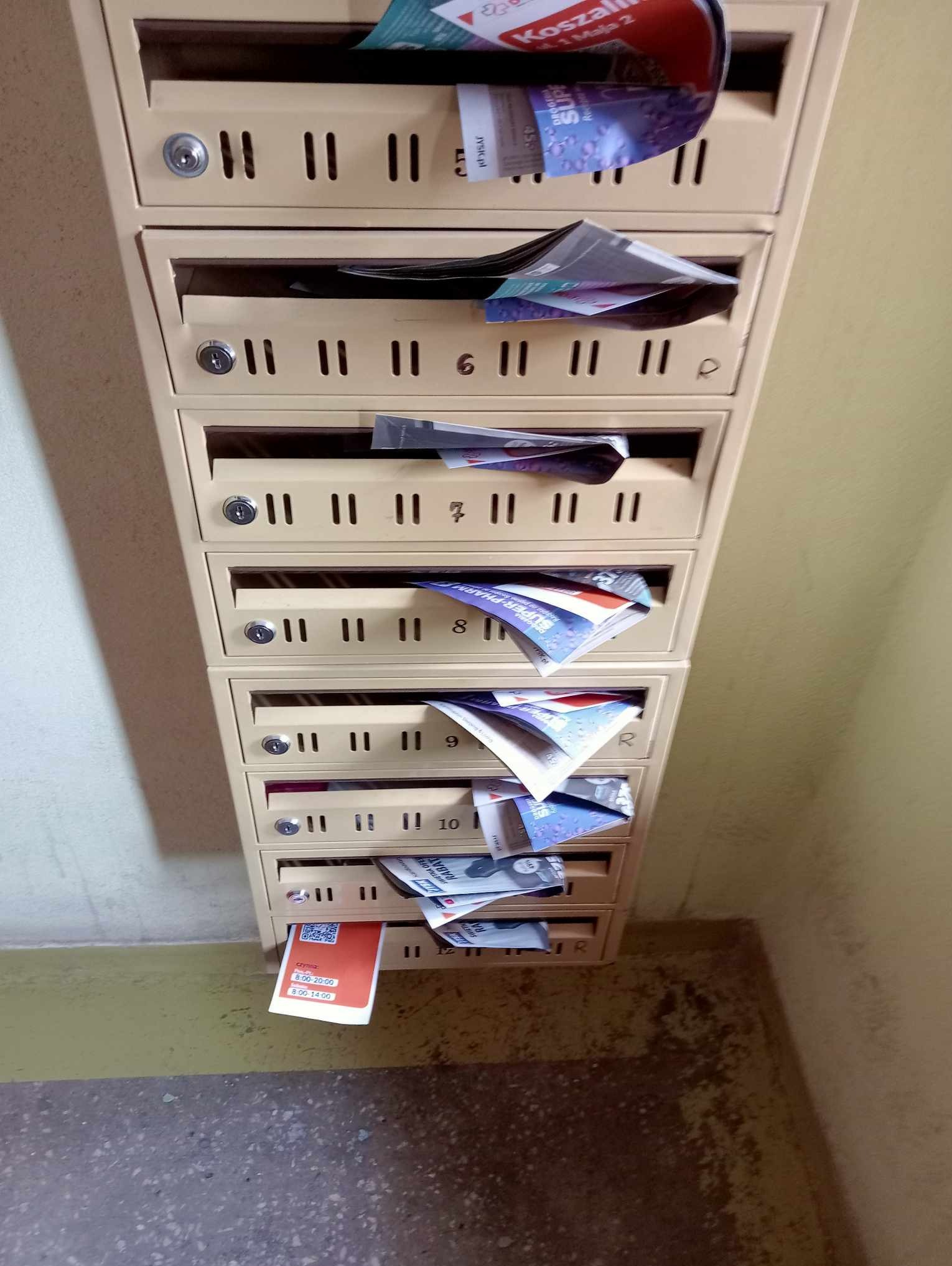 Skrzynki pocztowe w bloku mieszkalnym wypełnione ulotkami reklamowymi i informacyjnymi, niektóre wystają na zewnątrz.