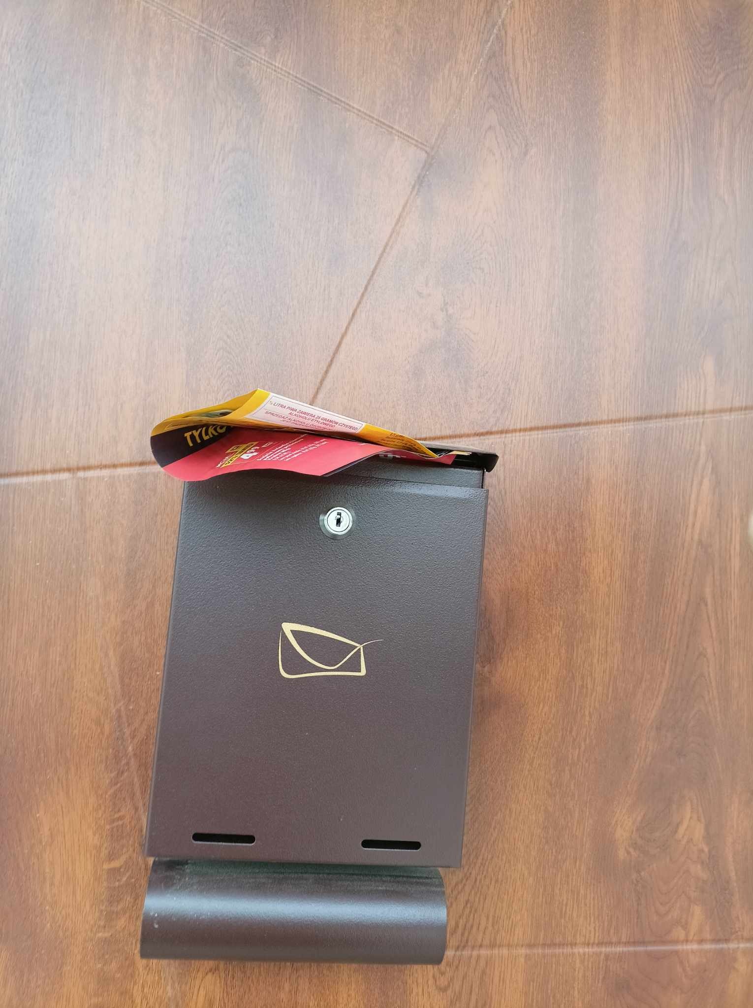Brązowa skrzynka pocztowa z wrzuconą ulotką reklamową firmy TYLKO, umieszczona na tle drewnianej ściany.