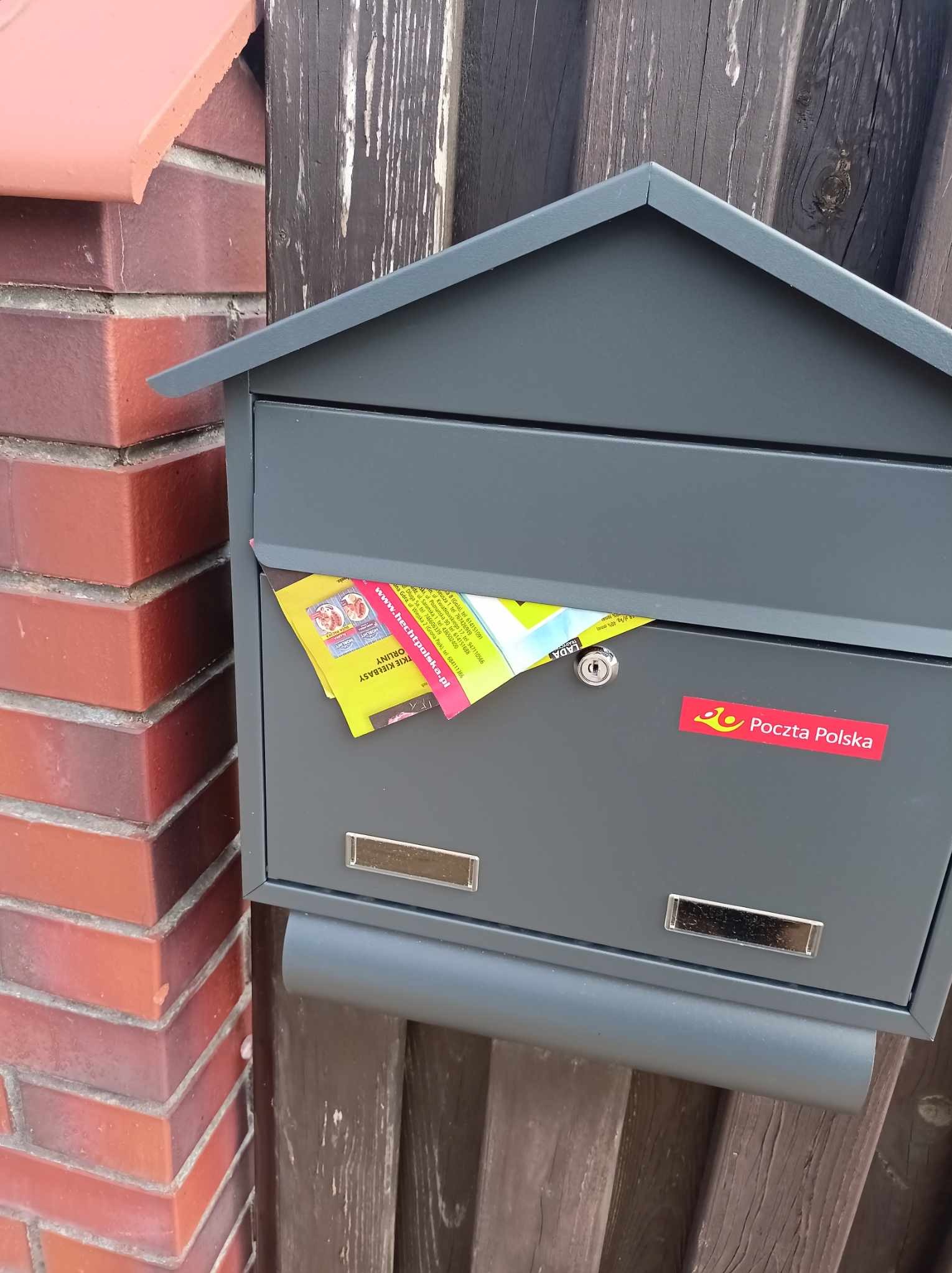 Szara skrzynka pocztowa z włożoną ulotką reklamową firmy z Koszalina, naklejka Poczty Polskiej na tle ceglanego muru i drewnianego płotu.