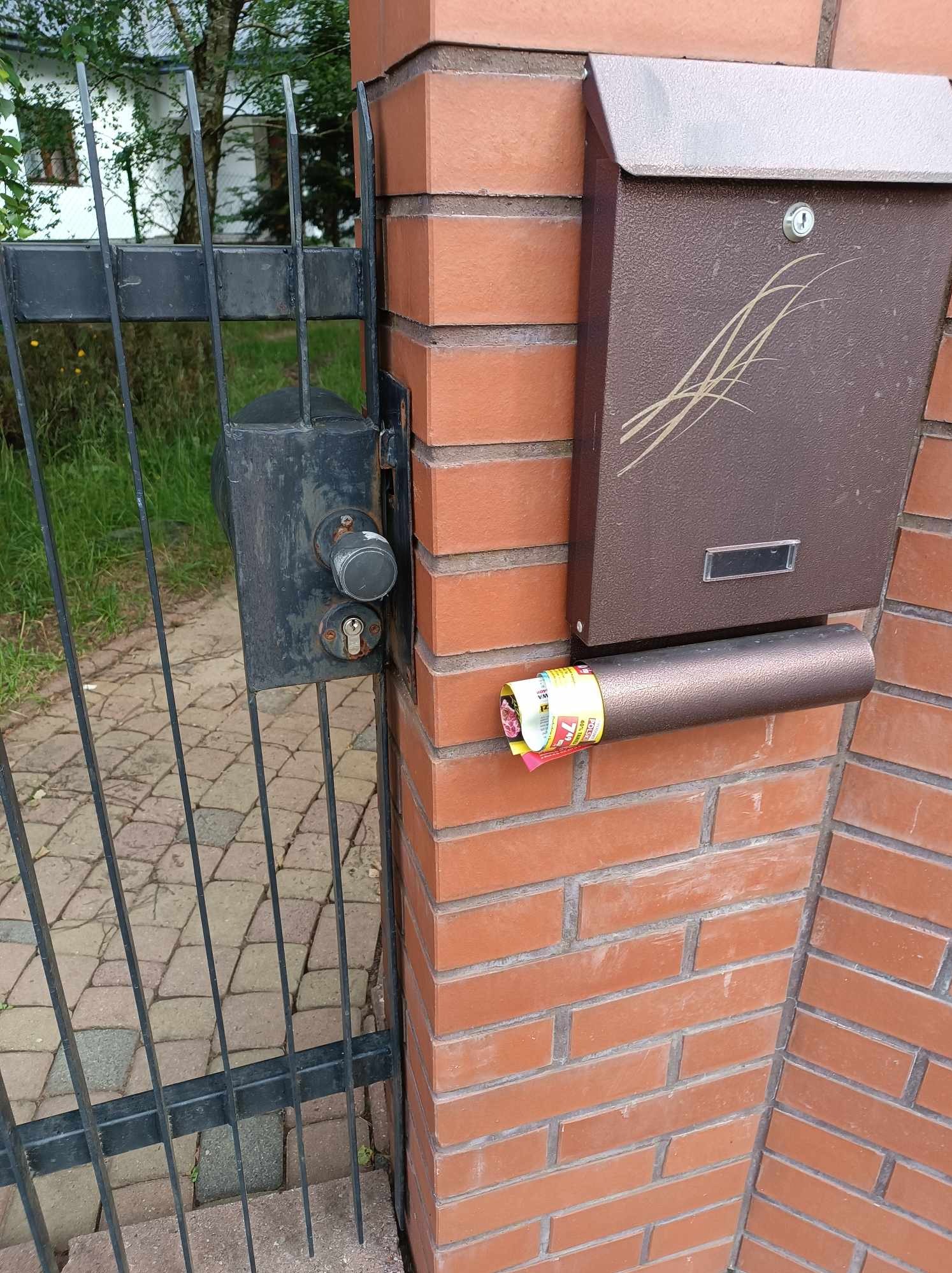 Brązowa skrzynka pocztowa z wsuwanym rulonem ulotek na ceglanym słupku ogrodzenia z czarną metalową furtką