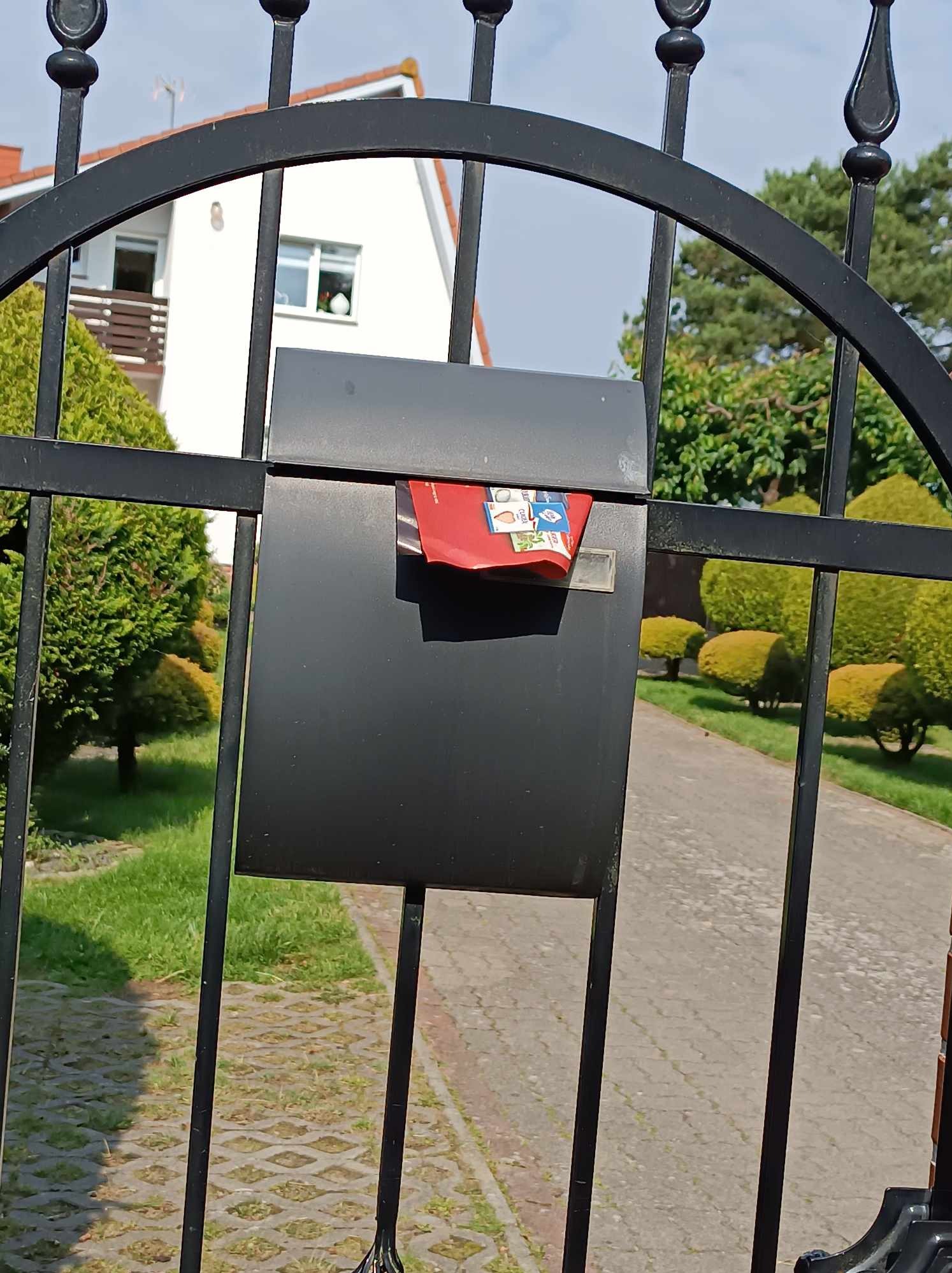 Czarna skrzynka pocztowa zamontowana na metalowym ogrodzeniu, wystają z niej czerwone ulotki, w tle dom z ogrodem.