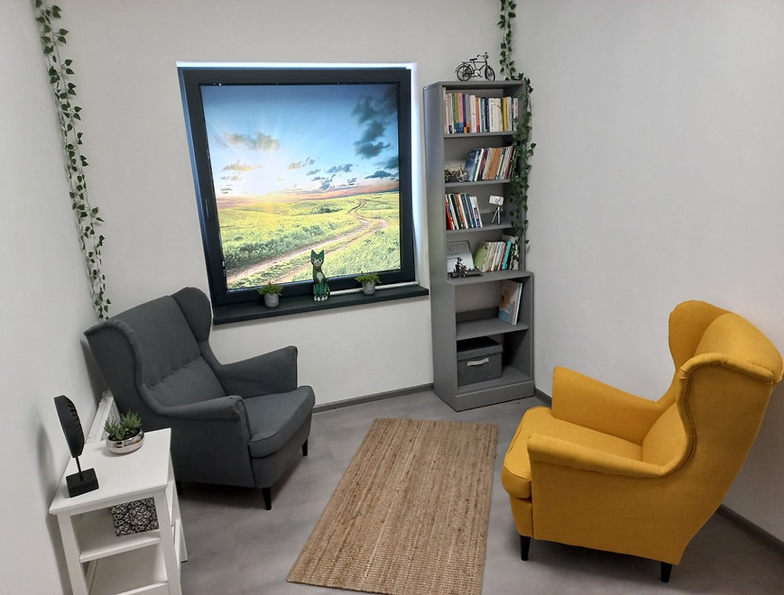 Pokój terapeutyczny z szarym i żółtym fotelem uszak, regałem z książkami, oknem z widokiem na krajobraz i dekoracjami roślinnymi.