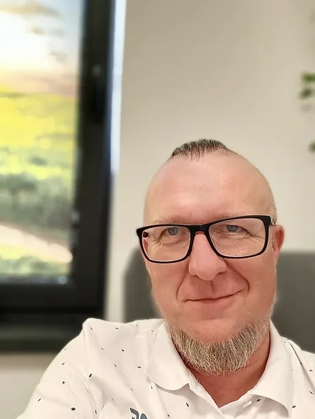 Portret mężczyzny w okularach i koszuli w kropki, z brodą, uśmiechającego się do kamery, w tle okno z widokiem na zachód słońca.