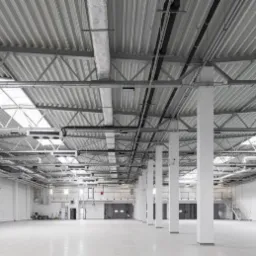 Wnętrze hali przemysłowej z widoczną konstrukcją stalową dachu, systemem wentylacyjnym i białymi słupami nośnymi. Podłoga jasna, gładka, prawdopodobnie epoksydowa. W tle widoczne bramy wjazdowe.