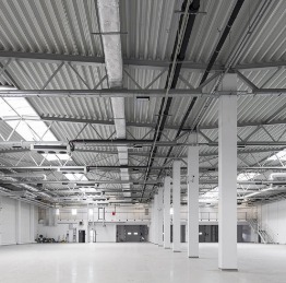 Wnętrze hali przemysłowej z widoczną konstrukcją stalową dachu, systemem wentylacyjnym i białymi słupami nośnymi. Podłoga jasna, gładka, prawdopodobnie epoksydowa. W tle widoczne bramy wjazdowe.