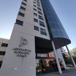 NEWPOINT FINANCE SPÓŁKA Z OGRANICZONĄ ODPOWIEDZIALNOŚCIĄ SPÓŁKA KOMANDYTOWA - Budynek biurowy Jesionowa Business Point z charakterystyczną szklaną fasadą, widziany z dołu pod kątem, w słoneczny dzień. Widoczne logo firmy na elewacji.