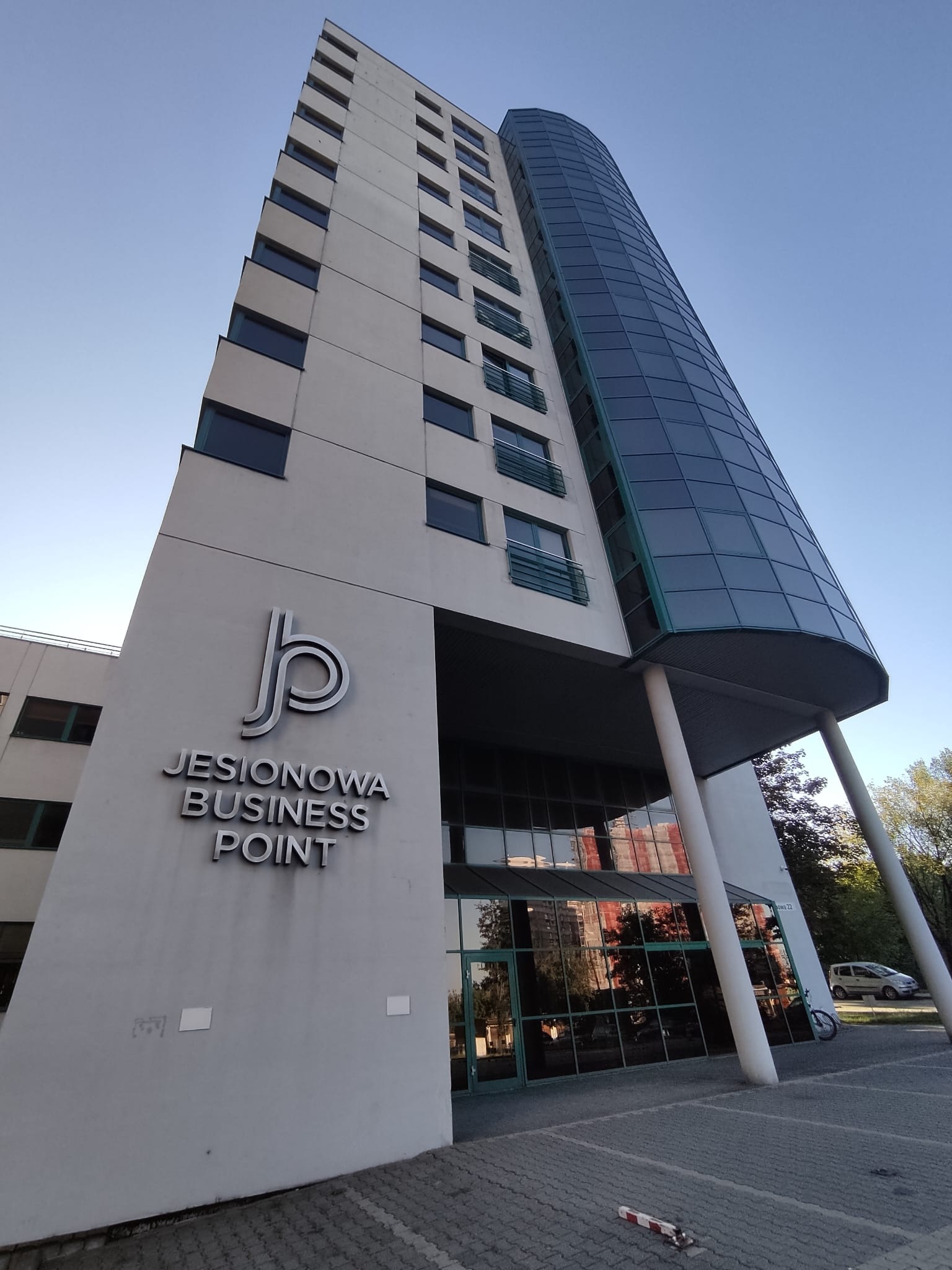 Budynek biurowy Jesionowa Business Point z charakterystyczną szklaną fasadą, widziany z dołu pod kątem, w słoneczny dzień. Widoczne logo firmy na elewacji.