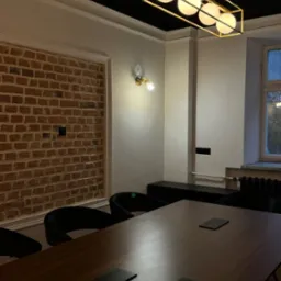 Nowoczesna sala konferencyjna z długim drewnianym stołem, czarnymi krzesłami, ceglaną ścianą i designerską lampą sufitową z czterema okrągłymi żar&oacute;wkami.