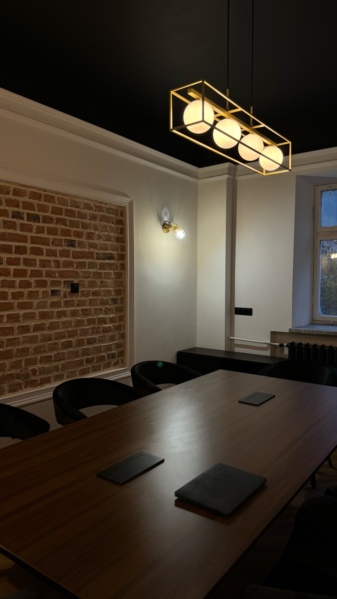 Nowoczesna sala konferencyjna z długim drewnianym stołem, czarnymi krzesłami, ceglaną ścianą i designerską lampą sufitową z czterema okrągłymi żarówkami.