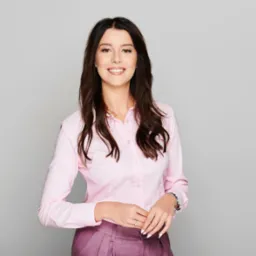 Portret uśmiechniętej kobiety w różowej koszuli i bordowej spódnicy, z długimi brązowymi włosami, na jasnoszarym tle.