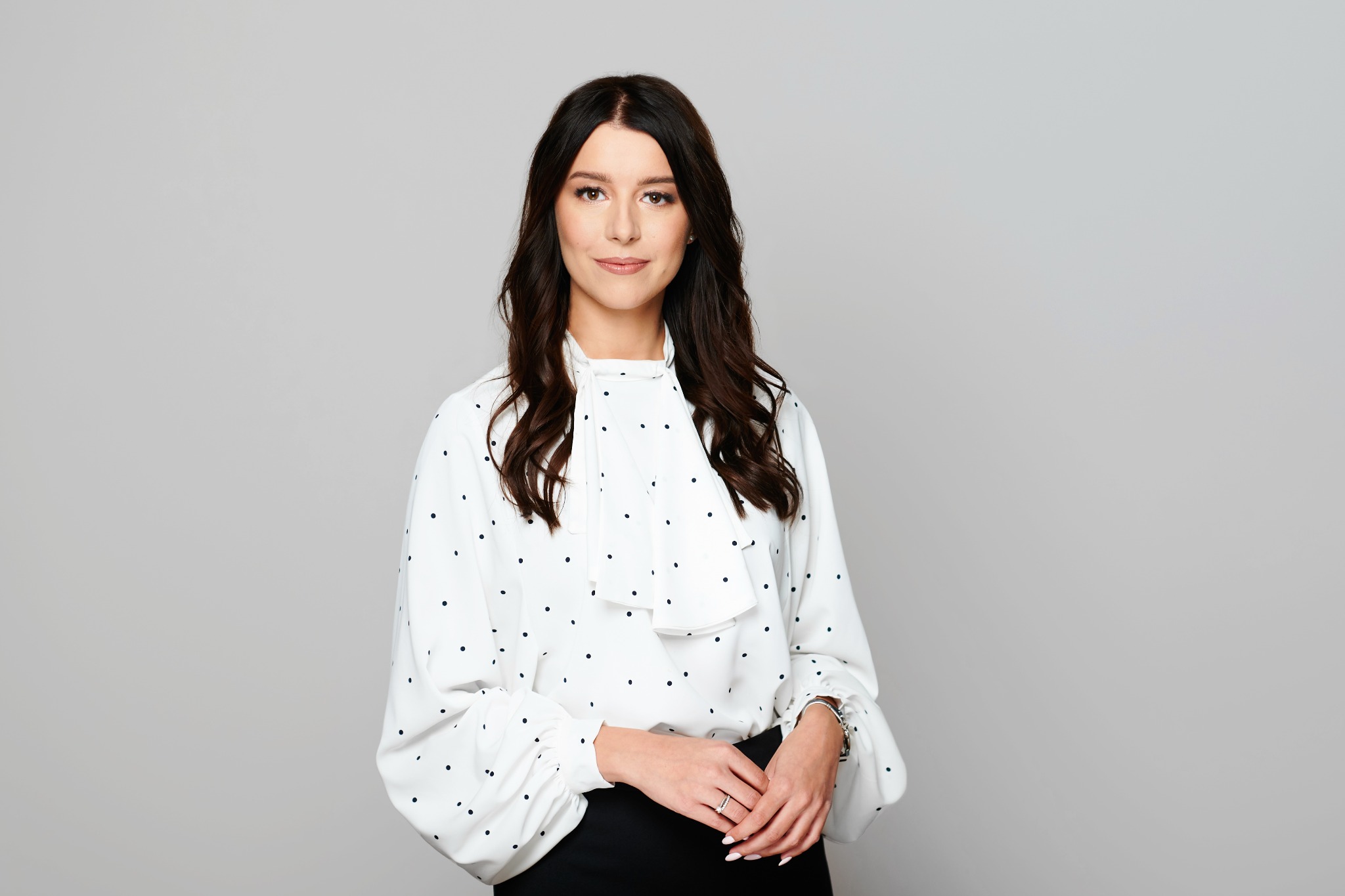 Portret kobiety w białej bluzce w czarne kropki i czarnej spódnicy, z delikatnym uśmiechem, stojącej na jasnoszarym tle.