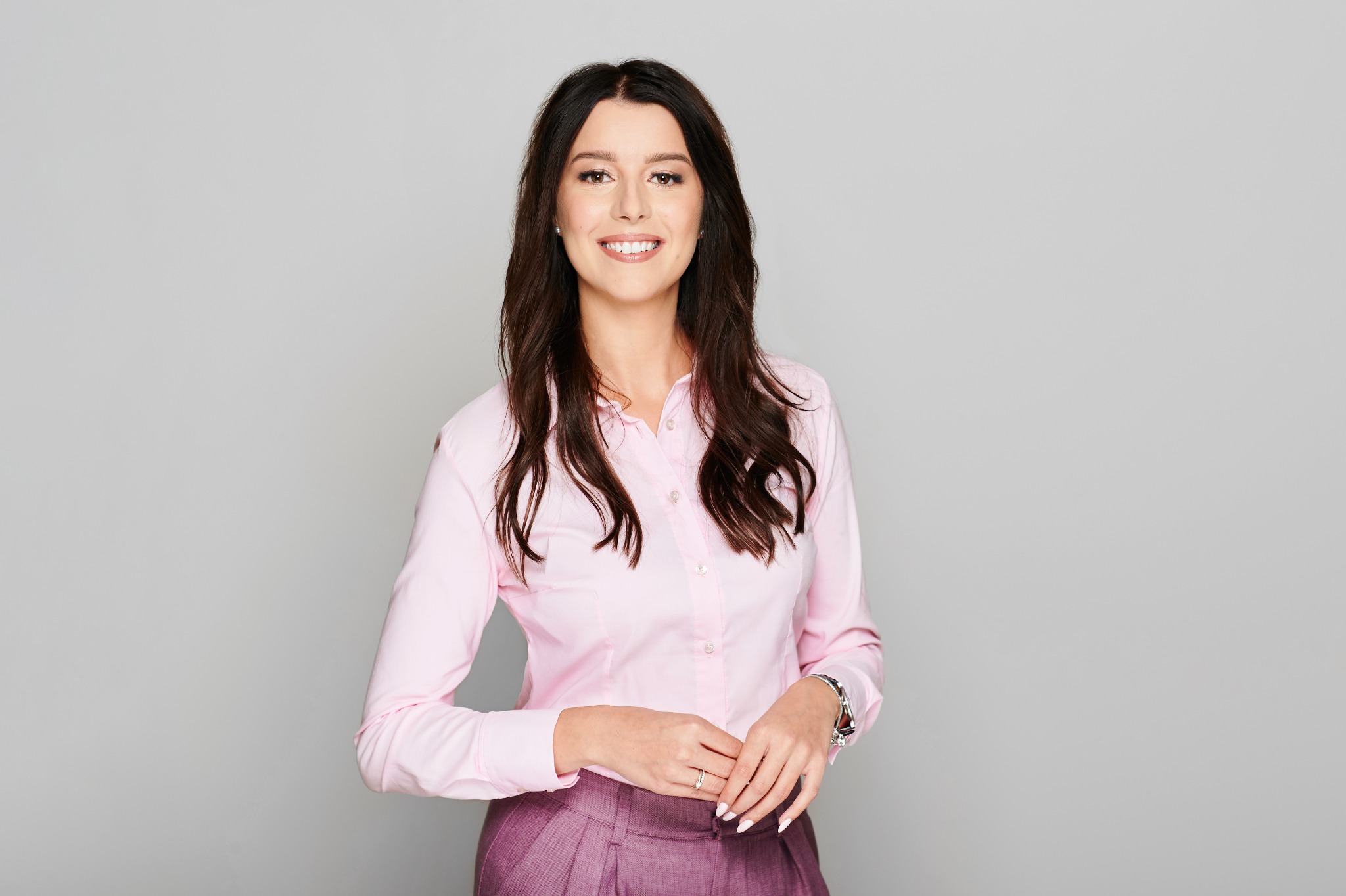 Portret uśmiechniętej kobiety w różowej koszuli i bordowej spódnicy, z długimi brązowymi włosami, na jasnoszarym tle.