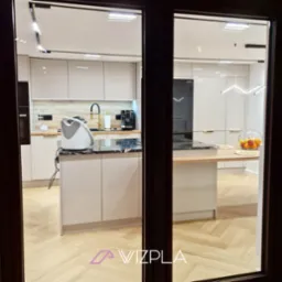 Nowoczesna kuchnia z wyspą, jasne lakierowane fronty, marmurowy blat, parkiet w jodełkę, widok przez uchylone drzwi.