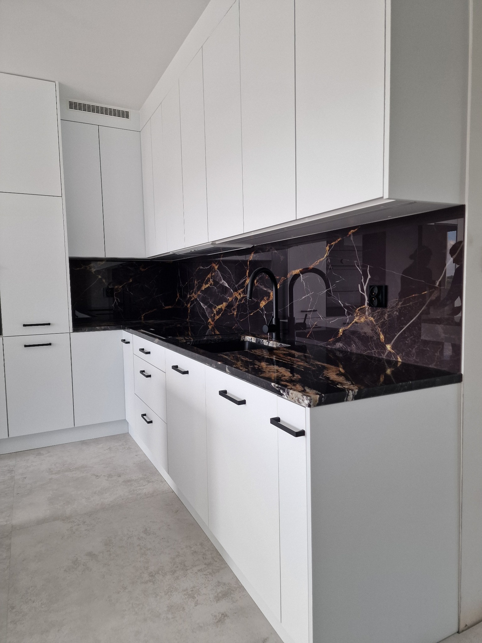 Biała zabudowa kuchenna z czarnym blatem i panelem ściennym imitującym marmur ze złotymi żyłami, czarna bateria kuchenna.