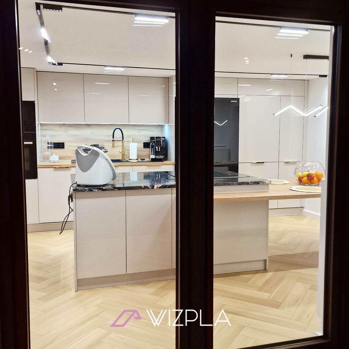 Nowoczesna kuchnia z wyspą, jasne lakierowane fronty, marmurowy blat, parkiet w jodełkę, widok przez uchylone drzwi.