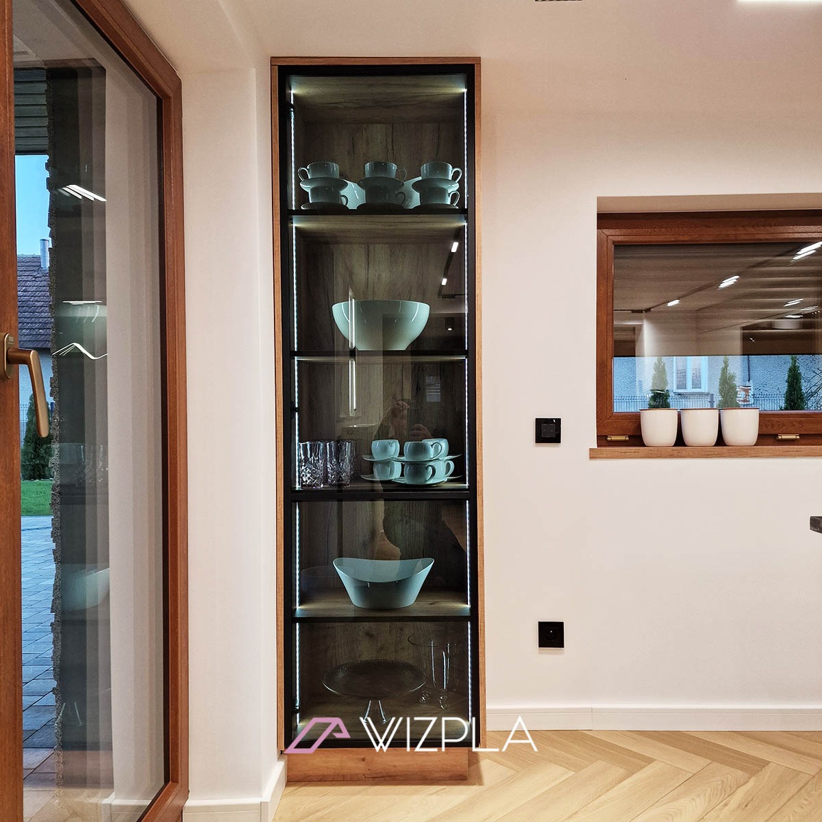 Wysoka, przeszklona witryna z podświetleniem LED, eksponująca zastawę stołową w nowoczesnej kuchni, widok z parkietem w jodełkę.