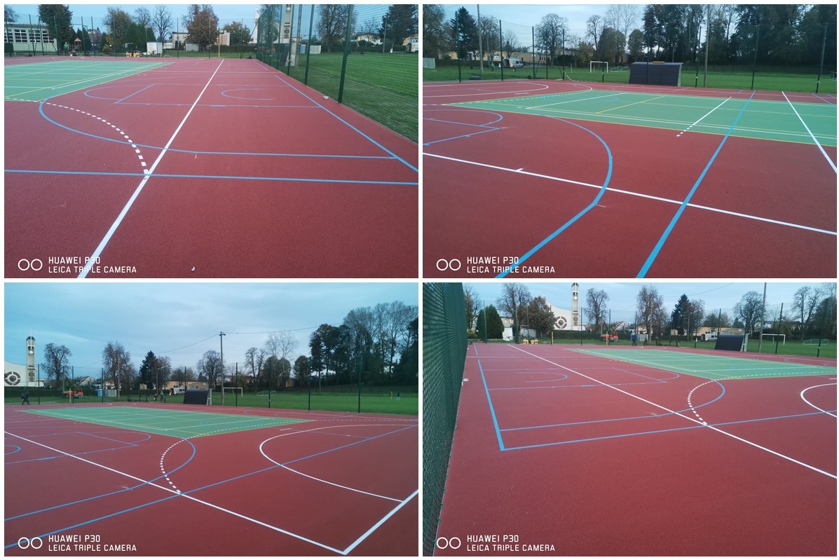 Wielofunkcyjne, nowo wybudowane boisko sportowe z nawierzchnią w kolorze czerwieni i zieleni, z wyraźnie oznaczonymi liniami w kolorze białym i niebieskim, otoczone zielenią i ogrodzeniem.